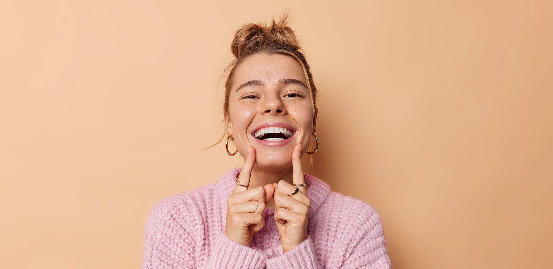 Positive young woman with combed hair points at her broad toothy smile feels very happy has white perfect teeth wears knitted sweater isolated over beige background. People and emotions concept
