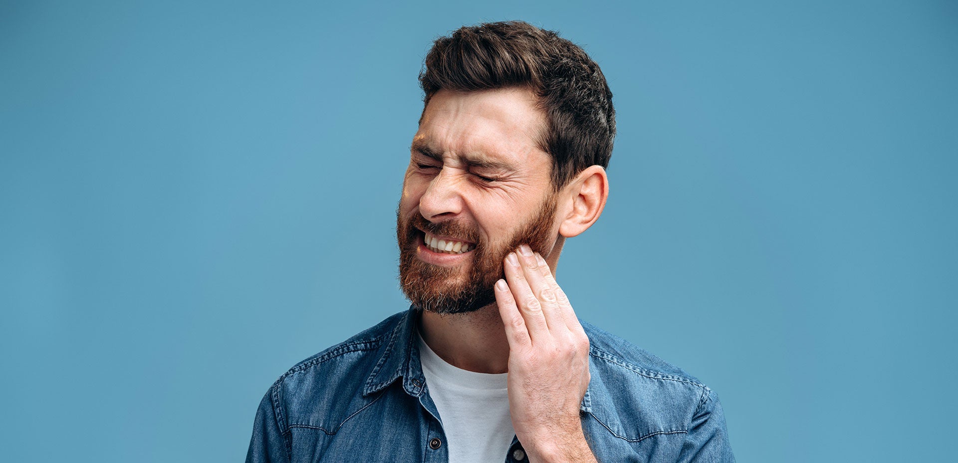 Dental problems. Portrait of unhealthy man pressing sore cheek, suffering acute toothache, periodontal disease, cavities or jaw pain. Indoor studio shot isolated on blue background